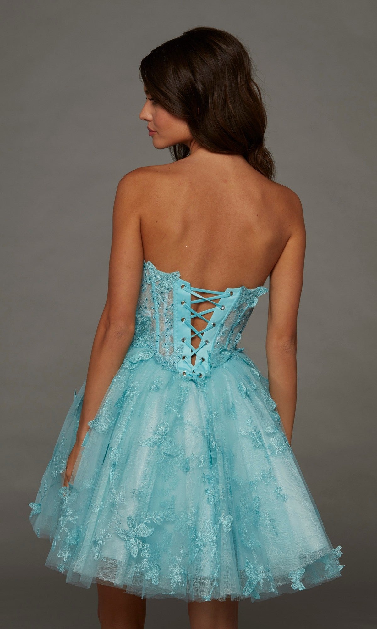A woman with brown hair wears the Short Homecoming Dress: Alyce 30097—a light blue, strapless dress with a corset-back and delicate lace and floral details—while standing against a plain gray background.